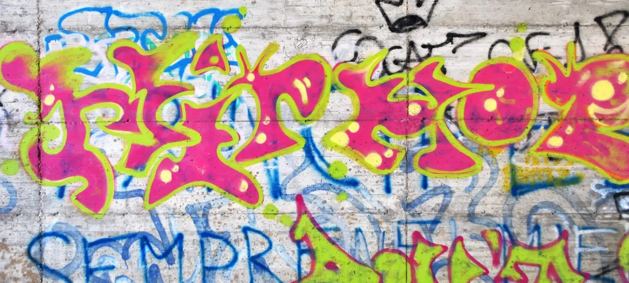 A close view of bright pink, green, and yellow graffiti painted on a rough concrete wall, layered over older blue and black markings.
